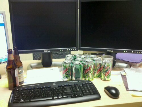 A scene of Mountain Dew cans and beer bottles on a desk with computer screens